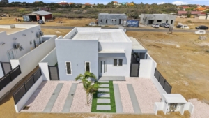 Elevated aerial view of Reina Sophia Model Luca Unit 20 in Aruba showing the white exterior, front landscaping, enclosed yard, and nearby properties.