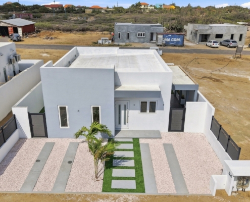 Elevated aerial view of Reina Sophia Model Luca Unit 20 in Aruba showing the white exterior, front landscaping, enclosed yard, and nearby properties.