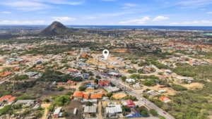 Wide aerial view around Reina Sophia Model Luca Unit 20 in Aruba showing residential streets, houses, vegetation, a prominent hill, and the sea in the distance.