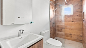 Reina Sophia Model Luca Unit 20 bathroom in Aruba with a white vanity, mirrored cabinet, toilet, glass shower enclosure, wood-look shower walls, and window.