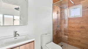 Reina Sophia Model Luca Unit 20 bathroom in Aruba with a white vanity, toilet, glass-enclosed shower, wood-look wall tiles, and small window.