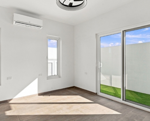 Reina Sophia Model Luca Unit 20 bedroom in Aruba with white walls, tiled floors, two windows, a wall-mounted air conditioner, and sliding glass doors opening toward a grassy outdoor area.