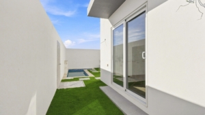 Outdoor courtyard at Reina Sophia Model Luca Unit 20 in Aruba with artificial grass, concrete stepping stones, white walls, glass doors, and a small pool in the background.