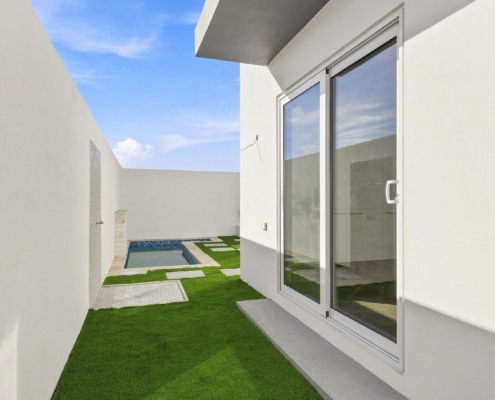 Outdoor courtyard at Reina Sophia Model Luca Unit 20 in Aruba with artificial grass, concrete stepping stones, white walls, glass doors, and a small pool in the background.