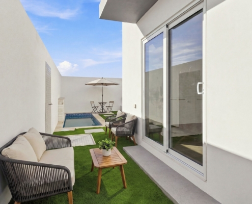 Outdoor courtyard at Reina Sophia Model Luca Unit 20 in Aruba with lounge seating, a wooden table, artificial grass, stepping stones, and a small pool.