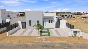 Aerial view of the front exterior of Reina Sophia Model Luca Unit 20 in Aruba showing the white house, enclosed yard, palm trees, gravel landscaping, and paved street.