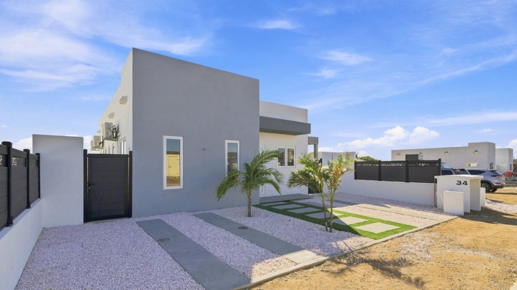 Front exterior of Reina Sophia Model Luca Unit 20 in Aruba with white walls, black fencing, palm trees, gravel landscaping, and concrete stepping paths.