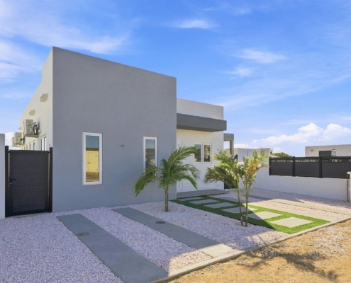 Front exterior of Reina Sophia Model Luca Unit 20 in Aruba with white walls, black fencing, palm trees, gravel landscaping, and concrete stepping paths.