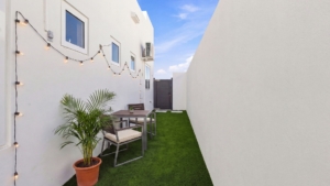 Side garden at Reina Sophia Model Luca Unit 20 in Aruba with artificial grass, a potted palm, outdoor dining furniture, string lights, and white walls.