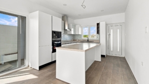 Reina Sophia Model Luca Unit 20 kitchen in Aruba with white cabinetry, a large island, built-in appliances, tiled backsplash, sink, and window.