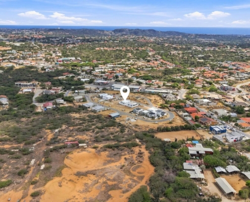 Aerial view from above the neighborhood around Reina Sophia Model Luca Unit 20 in Aruba showing residential buildings, roads, vegetation, and the coastline in the distance.