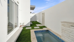 Private courtyard at Reina Sophia Model Luca Unit 20 in Aruba with a small rectangular pool, artificial grass, white walls, and large glass doors.