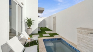 Pool courtyard at Reina Sophia Model Luca Unit 20 in Aruba with white lounge chairs, a small side table, a potted palm, artificial grass, and a rectangular pool.