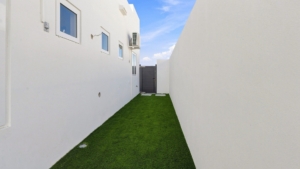 Side yard at Reina Sophia Model Luca Unit 20 in Aruba with artificial grass, white exterior walls, small windows, and a dark entrance gate.