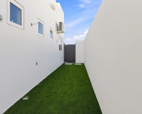 Side yard at Reina Sophia Model Luca Unit 20 in Aruba with artificial grass, white exterior walls, small windows, and a dark entrance gate.