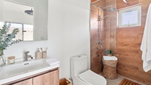 Staged Reina Sophia Model Luca Unit 20 bathroom in Aruba with a white sink vanity, toilet, glass shower, wood-look wall tiles, towels, basket, and decorative accessories.