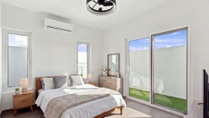 Staged Reina Sophia Model Luca Unit 20 bedroom in Aruba with a wooden bed, bedside tables, dresser, mirror, lamps, and sliding glass doors facing a grassy outdoor area.