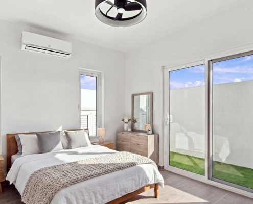 Staged Reina Sophia Model Luca Unit 20 bedroom in Aruba with a wooden bed, bedside tables, dresser, mirror, lamps, and sliding glass doors facing a grassy outdoor area.