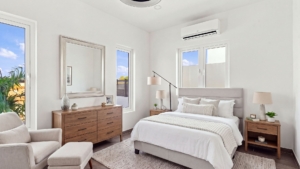 Staged Reina Sophia Model Luca Unit 20 bedroom in Aruba with a bed, wooden dresser, upholstered chair, lamps, mirror, and air conditioner.