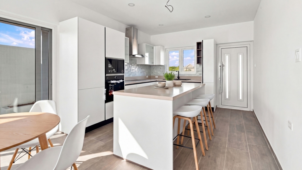 Staged Reina Sophia Model Luca Unit 20 kitchen in Aruba with white cabinets, a large island, bar stools, dining table, appliances, and backsplash.
