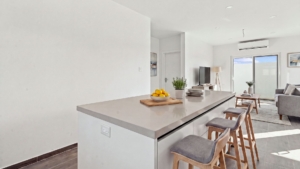Staged Reina Sophia Model Luca Unit 20 kitchen island in Aruba with a light countertop, wooden bar stools, fruit bowl, tableware, and living area beyond.