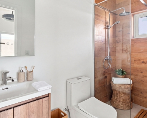 Staged Reina Sophia Model Luca Unit 21 bathroom in Aruba with a white sink vanity, toilet, glass shower, wood-look wall tiles, towels, basket, and decorative accessories.