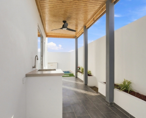 Covered outdoor area at Reina Sophia Model Luca Unit 22 in Aruba with a built-in counter and sink, tiled floor, wood ceiling, ceiling fan, and planted beds.
