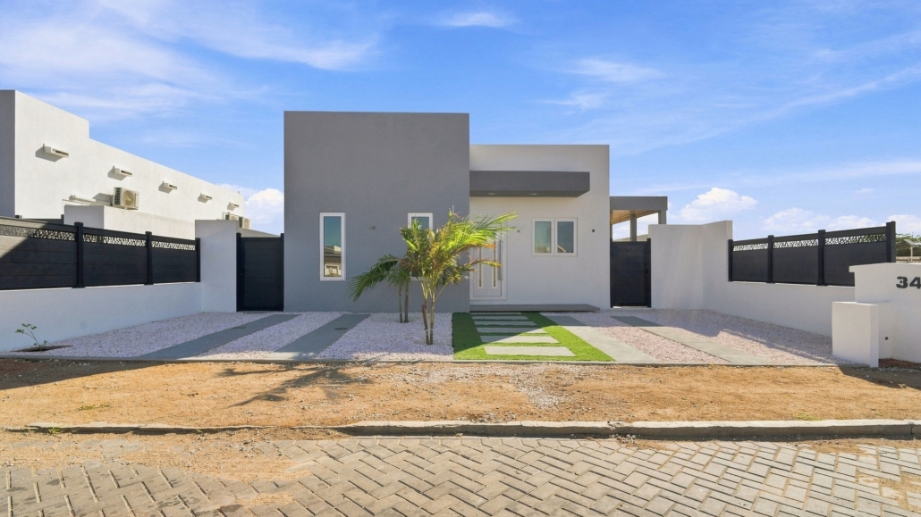 Aerial view of the front exterior of Reina Sophia Model Luca Unit 22 in Aruba showing the white house, enclosed yard, palm trees, gravel landscaping, and paved street.