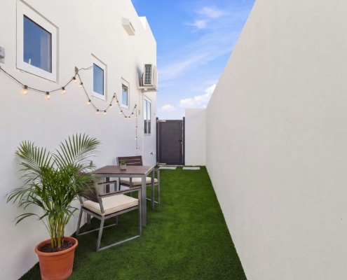 Side garden at Reina Sophia Model Luca Unit 22 in Aruba with artificial grass, a potted palm, outdoor dining furniture, string lights, and white walls.