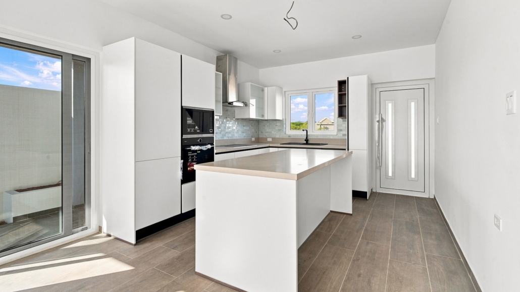Reina Sophia Model Luca Unit 24 kitchen in Aruba with white cabinetry, a large island, built-in appliances, tiled backsplash, sink, and window.