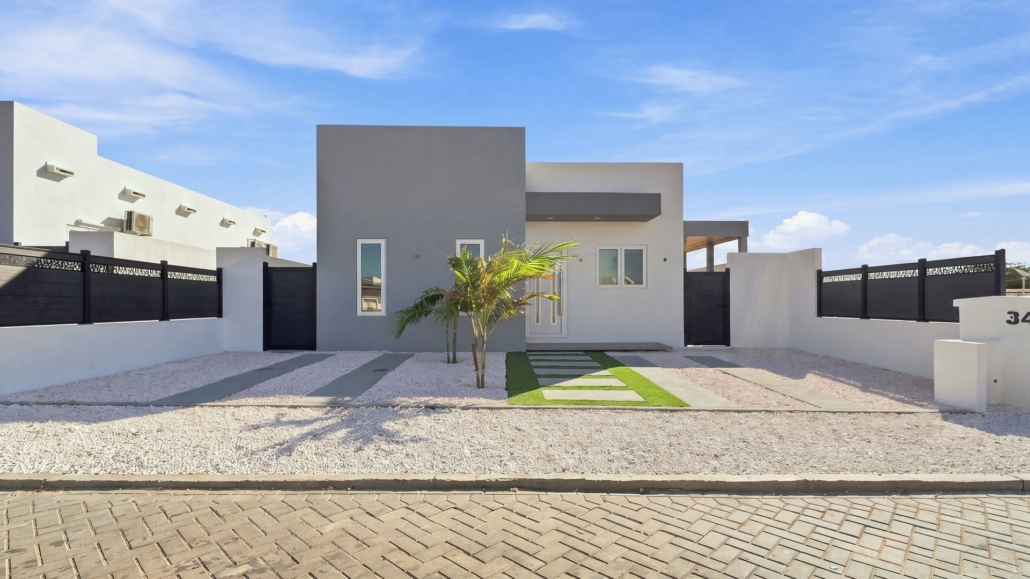 Modern front exterior of Reina Sophia Model Luca Unit 24 in Aruba with a white and gray facade, black fencing, palm trees, gravel landscaping, and stepping stones.