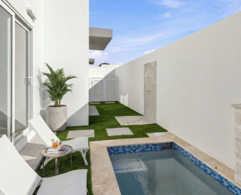 Pool courtyard at Reina Sophia Model Luca Unit 24 in Aruba with white lounge chairs, a small side table, a potted palm, artificial grass, and a rectangular pool.