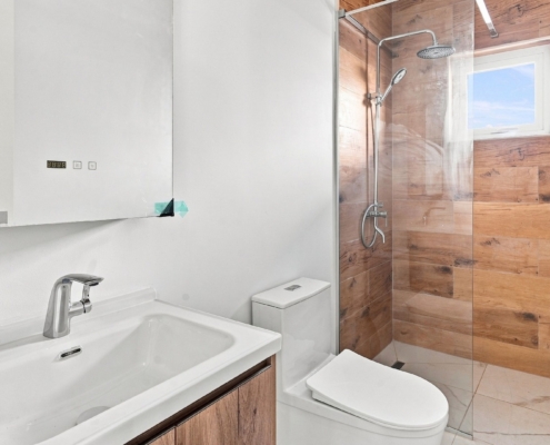 Reina Sophia Model Luca Unit 25 bathroom in Aruba with a white vanity, mirrored cabinet, toilet, glass shower enclosure, wood-look shower walls, and window.