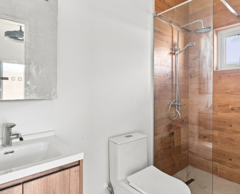 Bathroom with wood-look shower walls at Luca Unit 32 Aruba Reina Sophia Model Luca Unit 32 bathroom in Aruba with a white vanity, toilet, glass-enclosed shower, wood-look wall tiles, and small window.