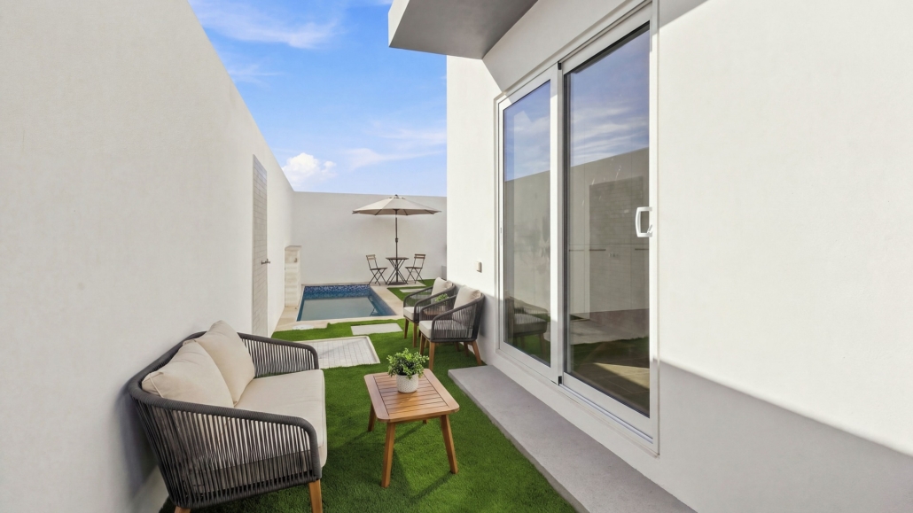 Outdoor courtyard at Reina Sophia Model Luca Unit 32 in Aruba with lounge seating, a wooden table, artificial grass, stepping stones, and a small pool.