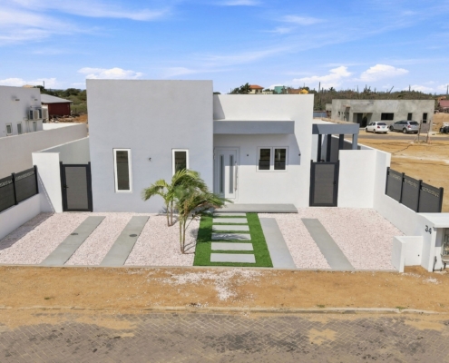 Reina Sophia Model Luca Unit 32 Front Exterior Aerial View in Aruba Aerial view of the front exterior of Reina Sophia Model Luca Unit 32 in Aruba showing the white house, enclosed yard, palm trees, gravel landscaping, and paved street.