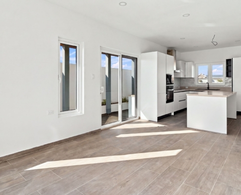 Open interior with kitchen view at Luca Unit 32 Aruba Reina Sophia Model Luca Unit 32 open interior in Aruba showing the kitchen island, sliding glass doors, windows, tiled floor, and white walls.
