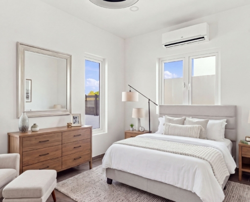 Staged bedroom at Reina Sophia Model Luca Unit 32 Aruba Staged Reina Sophia Model Luca Unit 32 bedroom in Aruba with a bed, wooden dresser, upholstered chair, lamps, mirror, and air conditioner.