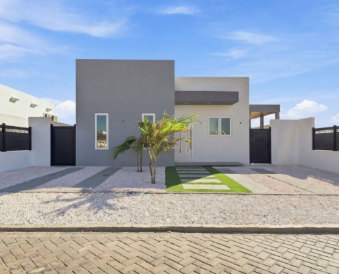 Reina Sophia Model Luca Unit 36 Modern Front Exterior in Aruba Modern front exterior of Reina Sophia Model Luca Unit 36 in Aruba with a white and gray facade, black fencing, palm trees, gravel landscaping, and stepping stones.
